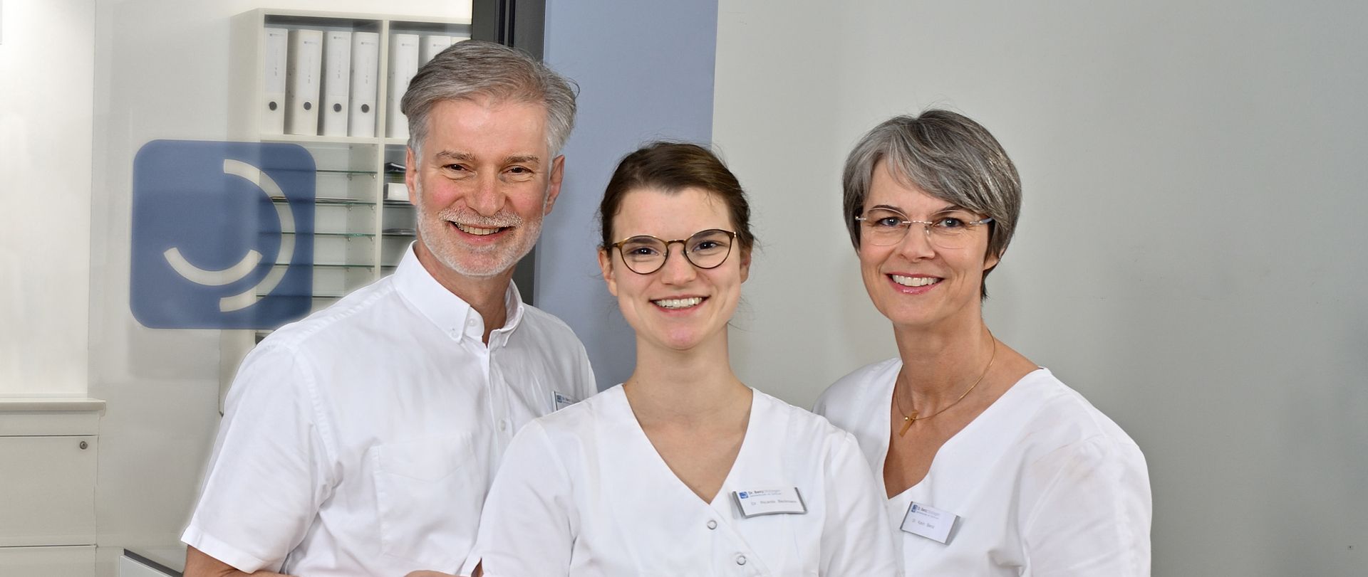 Herr Dr. med. dent. Jürgen Benz, Frau Dr. med. dent. Karin Benz und Frau Dr. med. dent. Ricarda Beckmann vor einer weißen Wand mit blauem Streifen